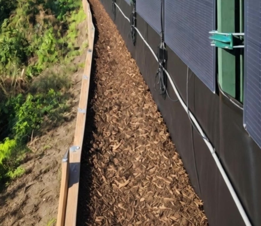 Modules solaires flexibles maintenus par des supports sur des poteaux de cl&ocirc;ture en acier vert, au-dessus d'une all&eacute;e en copeaux de bois